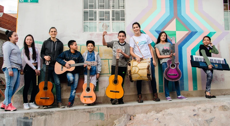 Grupo de personas diversas con instrumentos musicales parados frente a un colorido mural, todos unidos bajo el lema Al ComPAZ.