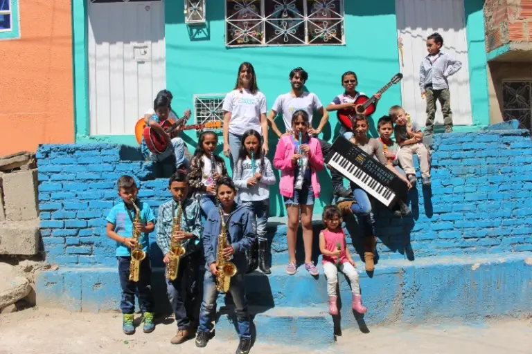 Grupo de niños y jóvenes con instrumentos musicales posando para una fotografía en las escaleras exteriores de un colorido edificio de Al ComPAZ.